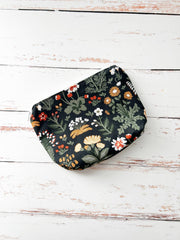 A small zippered pouch with a dark green floral and botanical print, displayed on a rustic white wooden plank surface.