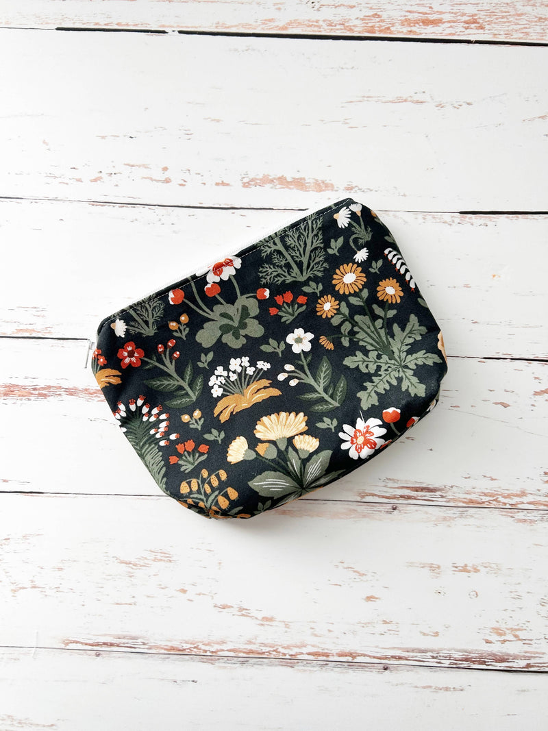 A small zippered pouch with a dark green floral and botanical print, displayed on a rustic white wooden plank surface.